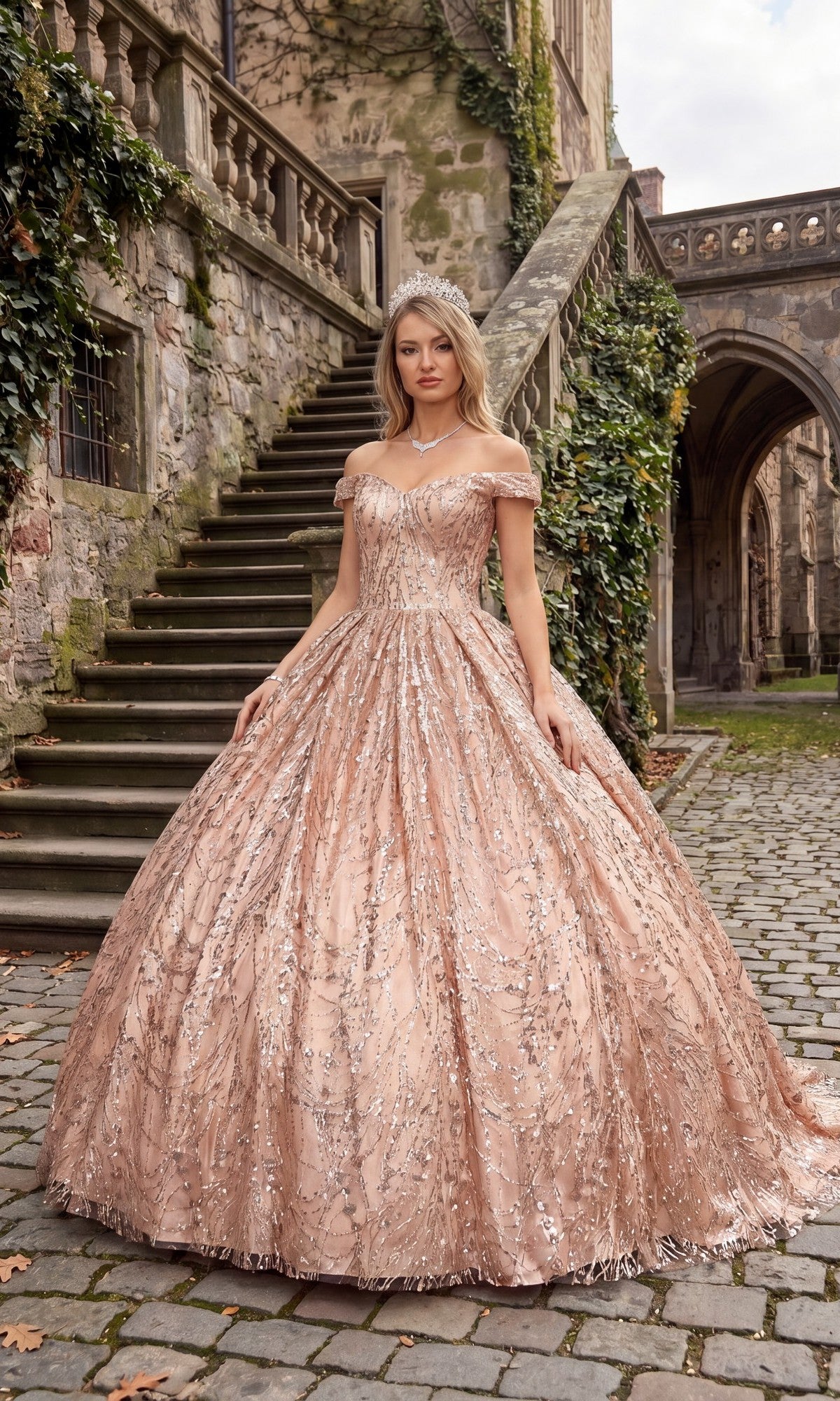 A woman in a sparkling, off-the-shoulder Quinceanera Dress BT9013 By Chicas in rose pink and a tiara stands on cobblestone near stone stairs and ivy-covered walls.